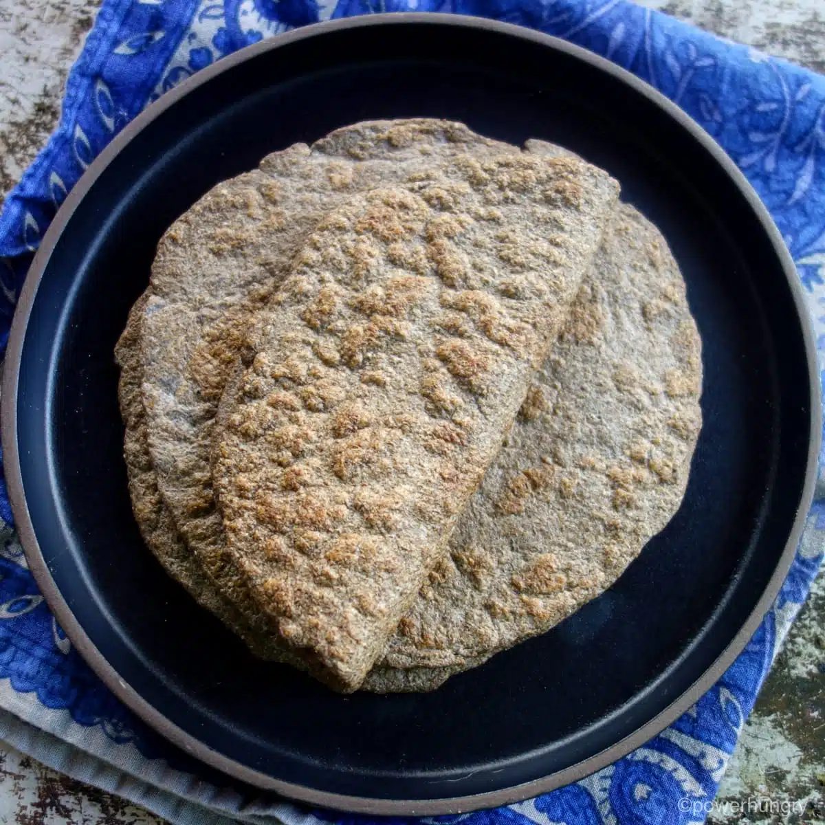 A stack of chia seed flatbreads on a black plate, positioned on top of a blue patterned napkin