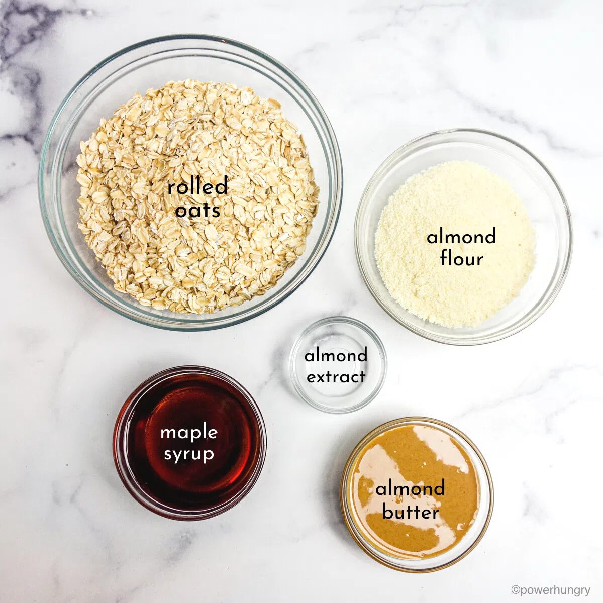 ingredients for almond granola, all in glass bowls, on a white marble surface