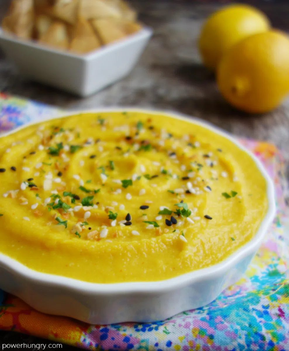 close up of lemon garlic split pea dip in a white bowl, with lemons and pita chips in the background
