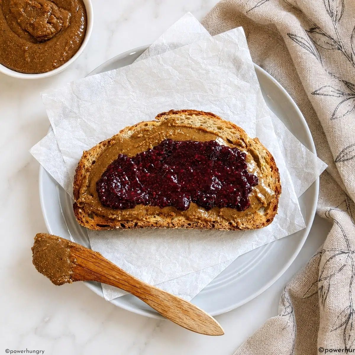 a piece of toast spread with roasted flaxseed butter and jam, set atop a pale grey plate