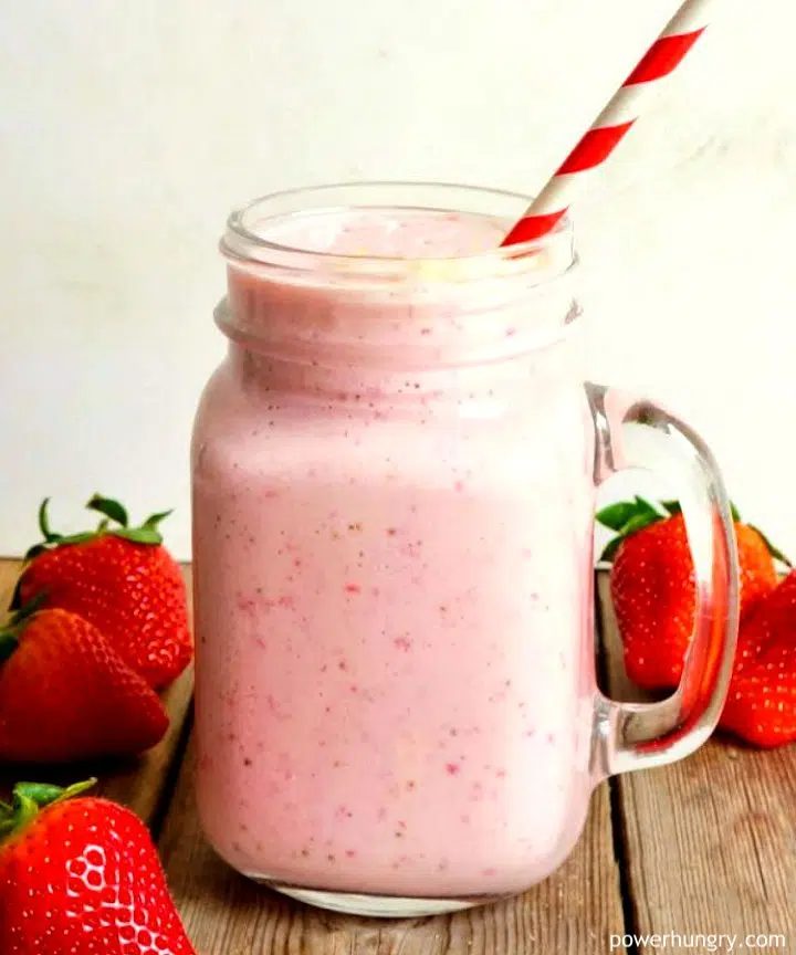 glass mug full of strawberry cashew smoothie with a red and white straw and several strawberries alongside