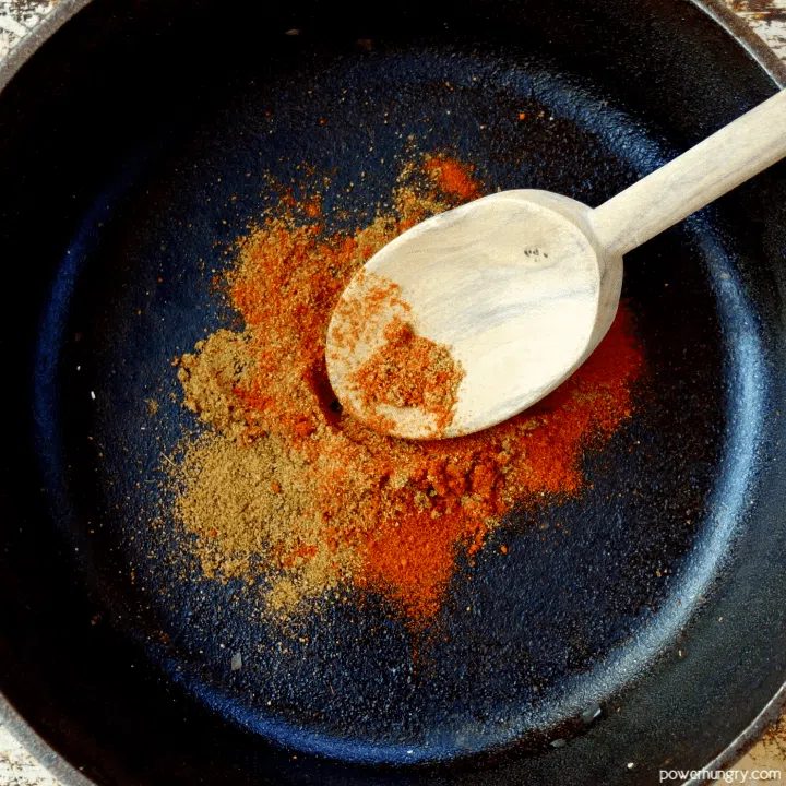 spices being toasted in a cast iron skillet, stirred with a wooden spoon