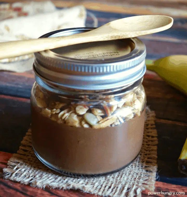 brownie batter breakfast oats, topped with granola, in a sealed jar, with a wooden spoon on top