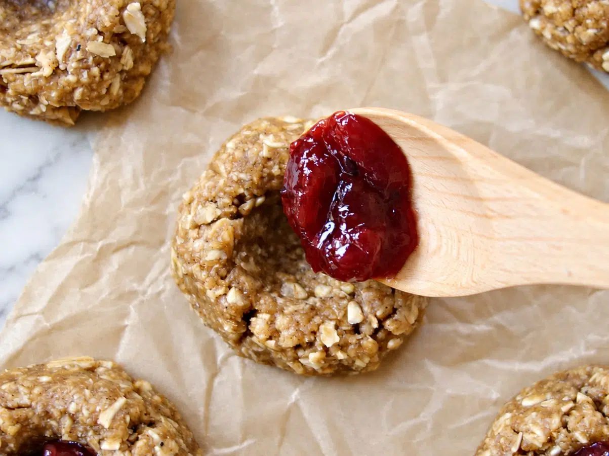 close-up of a spoonful of berry jam getting added to the center of a vegan oat flour thumbprint cookie