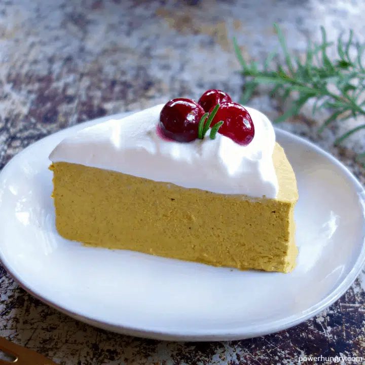 a slice of vegan hemp heart pumpkin cheesecake, topped with coconut whipped cream and sugared cranberries, on a white plate