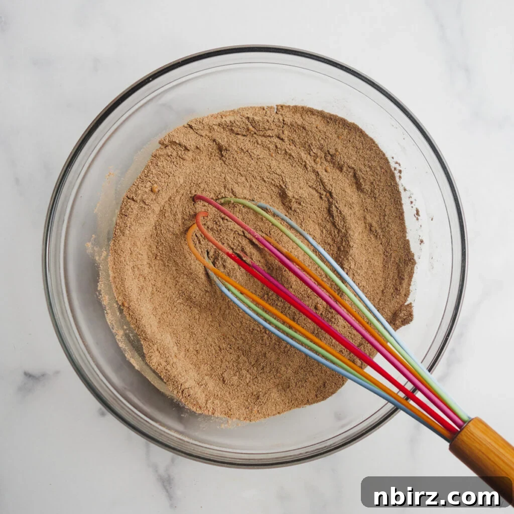 Indulgent Vegan Chocolate Buckwheat Cookies (Gluten-Free Oil-Free) 5 Dry chocolate buckwheat cookie ingredients in a glass bowl, whisked with a colorful whisk, showing a mix of light flour and dark cocoa powder.