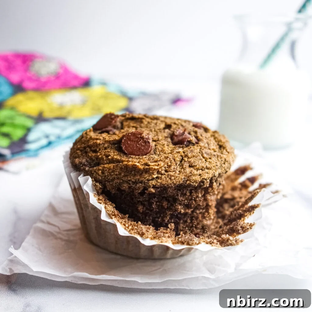 A single vegan gluten-free buckwheat muffin with visible chocolate chips, with its white paper liner partially peeled back, revealing its soft interior.