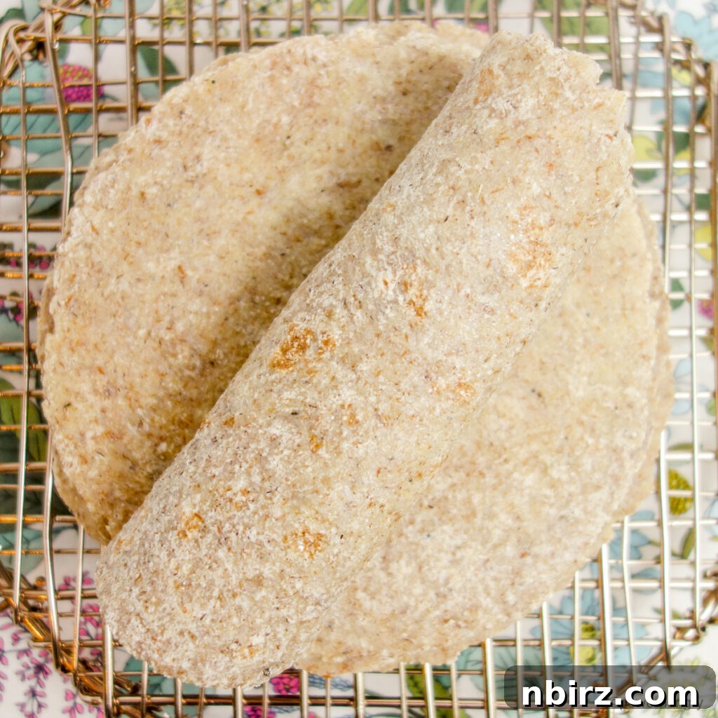 An overhead view of freshly cooked millet flax tortillas on a cooling rack, with one tortilla gently rolled to show its flexibility and texture.