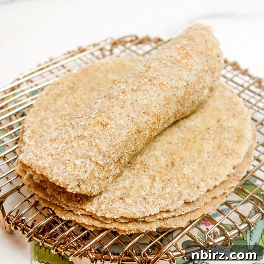 A single millet flax tortilla perfectly rolled into a tight cylinder, positioned atop a stack of more freshly made tortillas on a cooling rack, highlighting its flexibility.