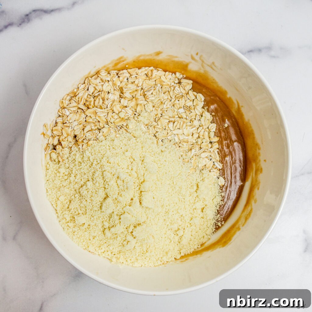 A moment captured as hearty rolled oats and delicate almond flour are gently poured into a mixing bowl, joining the rich, creamy wet ingredients for oil-free granola, poised to be combined into a cohesive, delicious mixture.