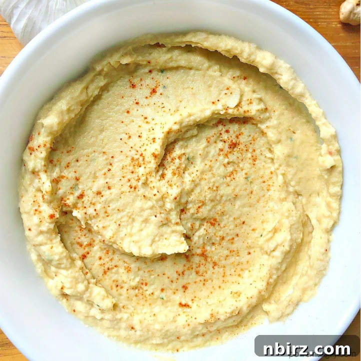 Creamy Oil-Free Hummus Without Tahini 2 Overhead shot of a white bowl filled with oil-free and tahini-free hummus, sprinkled with smoked paprika and fresh herbs.