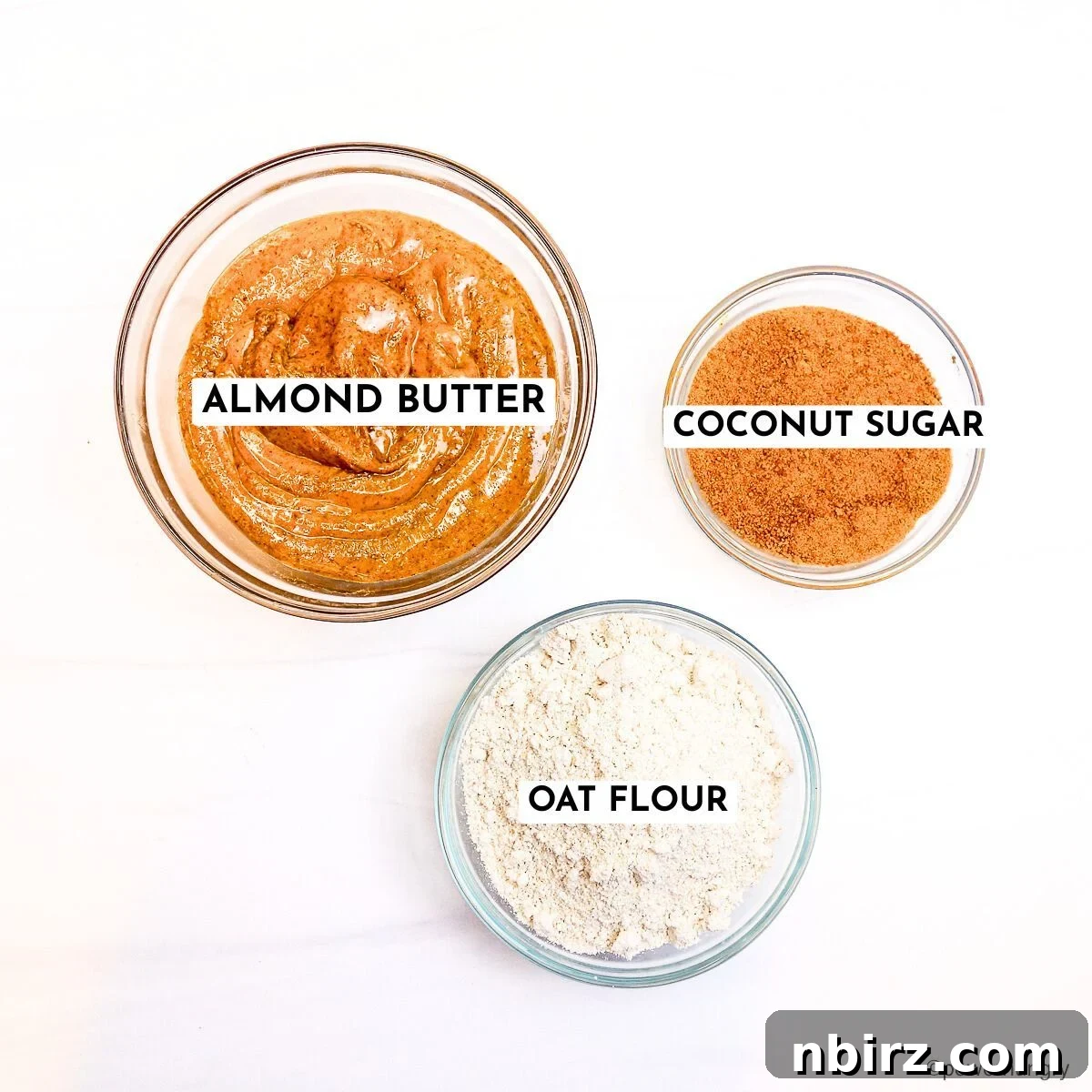 No-Fuss Almond Butter Oat Cookies 4 Glass bowls containing almond butter, oat flour, and coconut sugar on a marble surface, representing the 3 ingredients